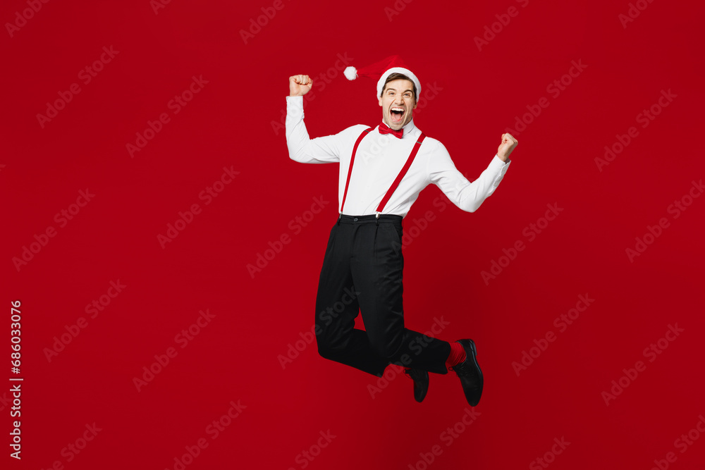 Full body merry young overjoyed excited man wear white shirt Santa hat posing jump high do winner gesture isolated on plain red background studio. Happy New Year Christmas celebration holiday concept. Full body merry young overjoyed excited man wear white shirt Santa hat posing jump high do winner gesture isolated on plain red background studio. Happy New Year Christmas celebration holiday concept.