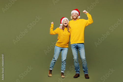 Full body merry young couple two friends man woman wear sweater Santa hat posing do winner gesture celebrate clench fists isolated on plain green background. Happy New Year Christmas holiday concept.