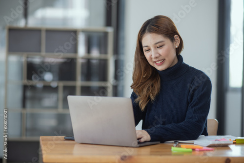 Happy young asian businesswoman with laptop sitting at workplace in modern office.