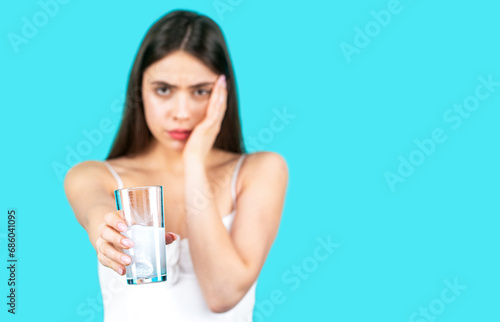 Woman taking pill against headache. Brunette taking a pill with a glass of water. Woman taking drugs to releave headache. Girl take some pills, holds glass of water, isolated on blue photo