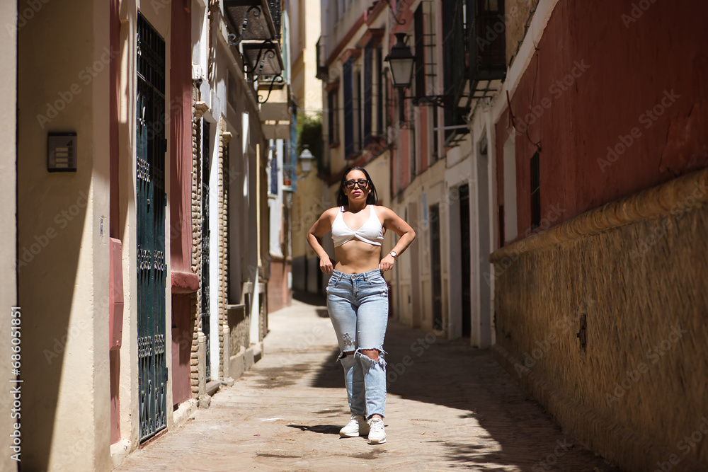 Young, beautiful, brunette and South American woman with white top, torn jeans and sunglasses, with her hands on her waist, receiving the sun's rays on a lonely street. Concept peace, tranquility.