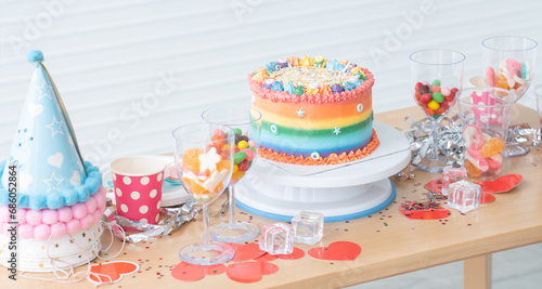 Cake on birthday with rainbow butter cream decorated with sugar candies  colorful sprinkles. Beautiful cake placed on stand with colorful candies  jelly in glasses and hats on table. Festive celebrate