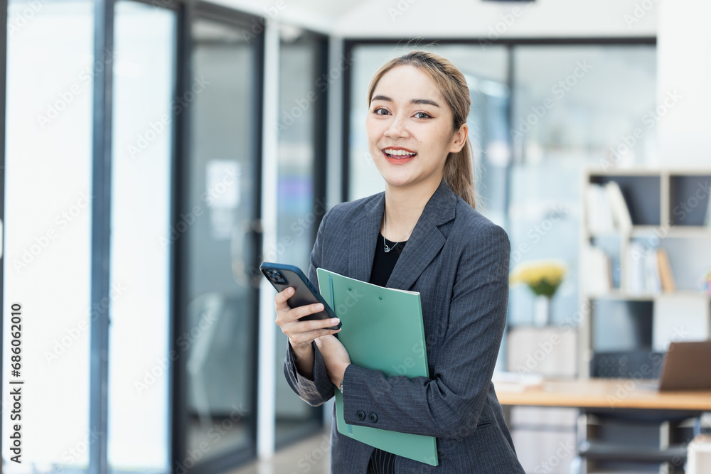 Asian businesswoman working on smartphone and financial document paper standing at desk in office workplace. Asian businesswoman working on smartphone and financial document paper standing at desk in office workplace.