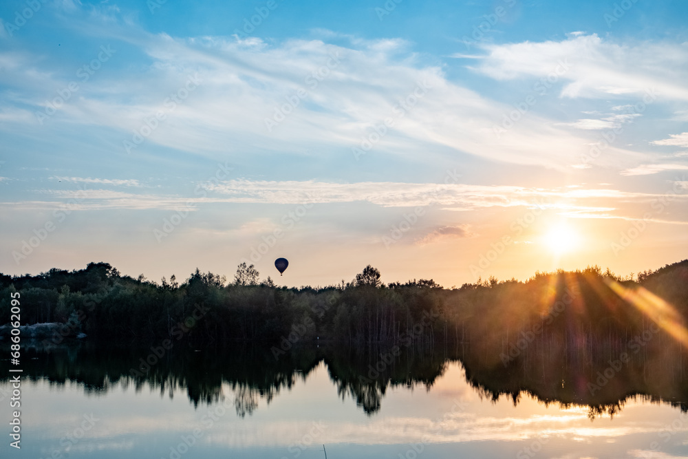 Captured at the moment when dawn breaks, this image presents a serene lake reflecting the awakening sky. The sun crests the horizon, scattering radiant beams and awakening the world with soft light. A