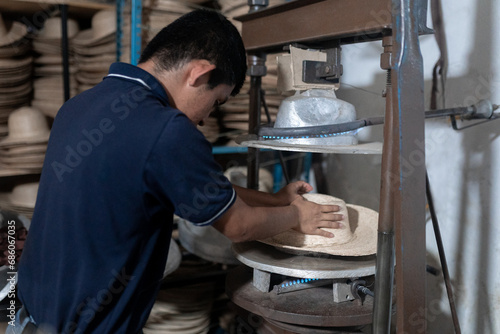 A young Hispanic man is shaping a natural fiber hat in a blocking machine