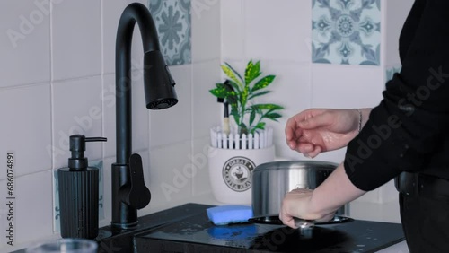 Medium shot of a young Woman wearing black clothes washing dishes (the Pot) with soap in black kitchen Sink under running water in slow motion. Shot with Sony FX3 in 4K photo