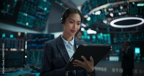 Portrait of a Beautiful Asian Female Working in an International Stock Exchange Hall: Specialist Wearing Headphones and Using Tablet Computer, Successful Trader Looking at Camera and Smiling photo