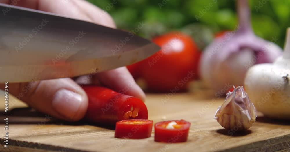 Extreme close-up of hands cutting a chilly pepper on a wooden board, various vegetables and herbs are laid out in the background. Extreme close-up of hands cutting a chilly pepper on a wooden board, various vegetables and herbs are laid out in the background.