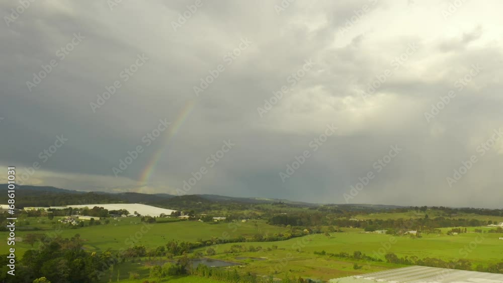 Aerial view pans gently to the left, showcasing a beautiful natural landscape under a colorful rainbow against a backdrop of thick clouds in the sky. Aerial view pans gently to the left, showcasing a beautiful natural landscape under a colorful rainbow against a backdrop of thick clouds in the sky.