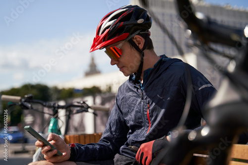Man in a bicycle helmet sits on a bench and looks at his phone, resting while cycling in the city