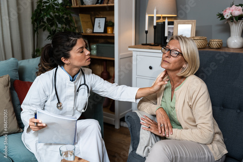 During a home visit to an elderly patient, a young endocrinologist doctor checks her thyroid gland by feeling her neck with her hands. Senior women endocrinology health care photo