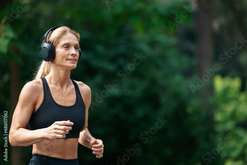 Athletic strong blonde runner woman in headphones jogging in park.