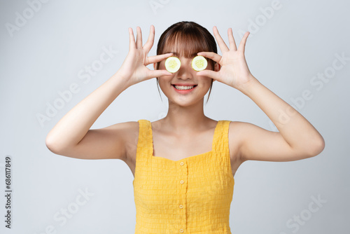 Beautiful and smiling young woman holding slice of cucumber in front of eyes on white background