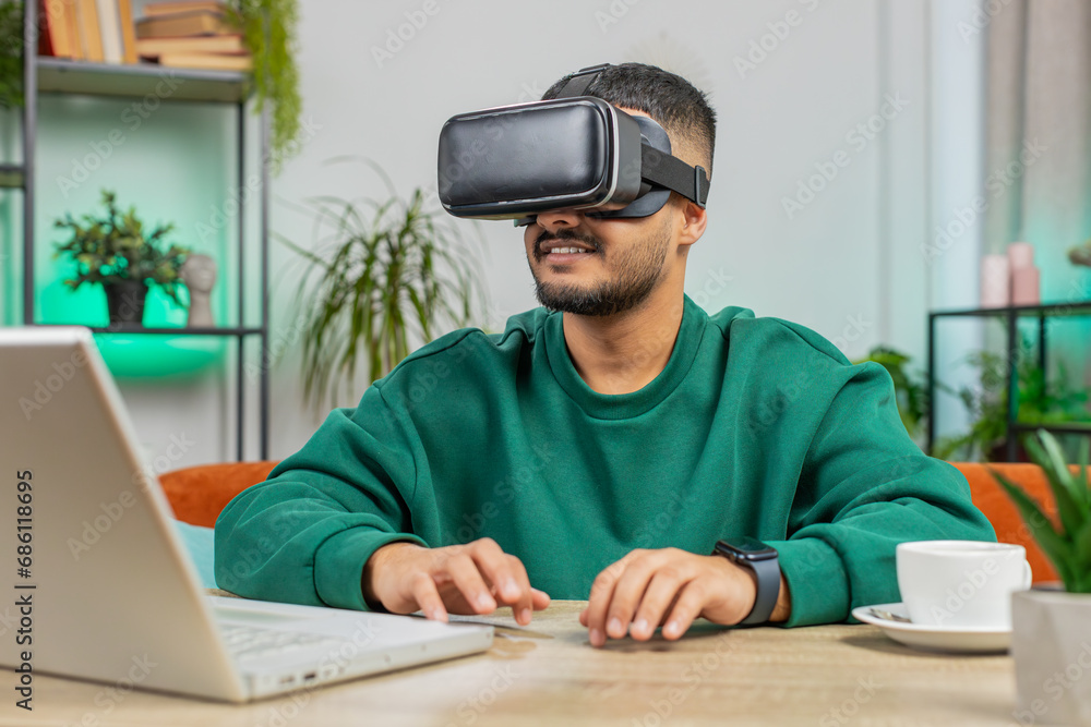 Indian man using virtual reality futuristic technology VR app headset helmet typing on virtual laptop keyboard, play simulation 3D 360 online video game at home office. Guy in goggles sitting at table Indian man using virtual reality futuristic technology VR app headset helmet typing on virtual laptop keyboard, play simulation 3D 360 online video game at home office. Guy in goggles sitting at table