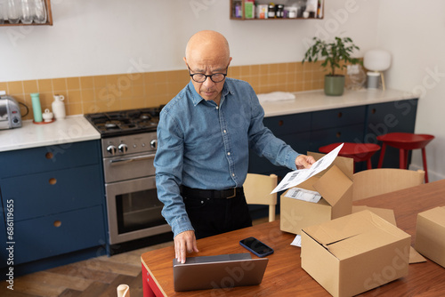 Senior male businessman packing orders into cardboard boxes to ship and using a laptop photo