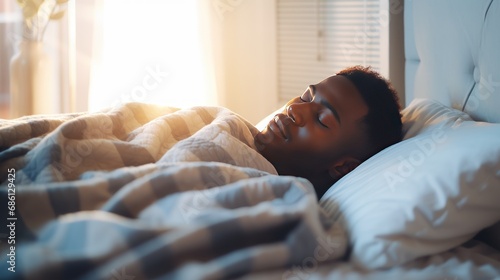 African-American man sleeps under warm plaid on soft bed at home closeup. Handsome black guy dreams lying on pillows in cozy bedroom. Young man naps comfortably in semi dark hotel room