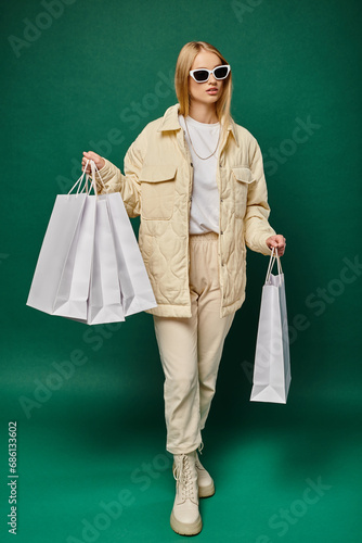 stylish blonde woman in winter wear and sunglasses walking with shopping bags on green, full length © LIGHTFIELD STUDIOS