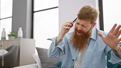 Irish redhead man, sitting in the living room, stressed about his conversation on his smartphone, fighting with frustration and despair photo