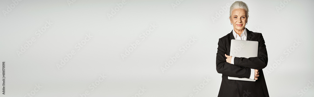 happy middle aged businesswoman in formal wear standing and holding laptop isolated on grey, banner happy middle aged businesswoman in formal wear standing and holding laptop isolated on grey, banner