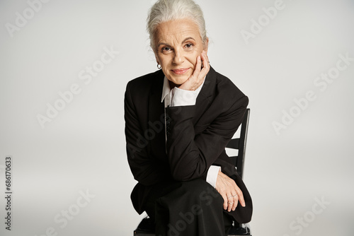 middle aged businesswoman in formal wear smiling while sitting on chair on grey backdrop