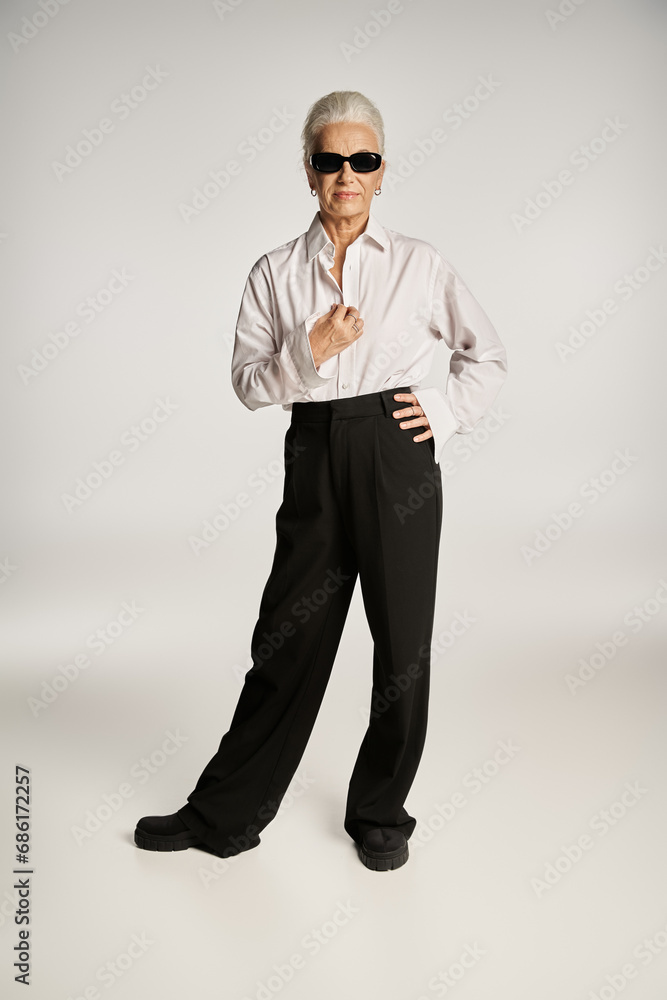 stylish middle aged woman in classy white shirt and sunglasses posing with hand on hip on grey stylish middle aged woman in classy white shirt and sunglasses posing with hand on hip on grey