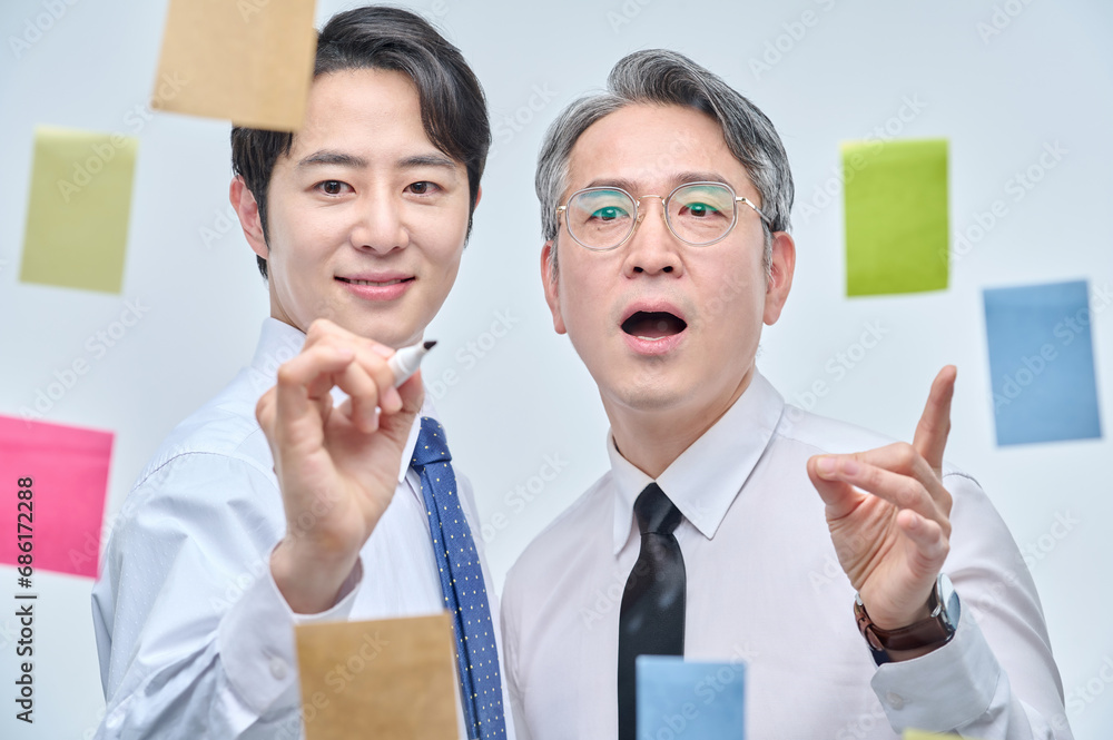 Two businessmen are standing on the glass wall of the office, looking at the attached paper and contemplating planning and strategy. Two businessmen are standing on the glass wall of the office, looking at the attached paper and contemplating planning and strategy.