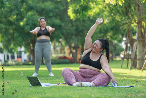 Asian female overweight in sportswear training exercise to lose weight on mat looking preview with laptop. woman plus size training online practice yoga in park. concept of healthy.
