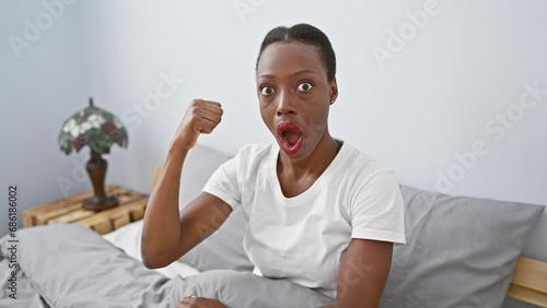 Amazed african american woman in bedroom, lying on bed with a surprised expression, giddily pointing, mouth wide open! photo