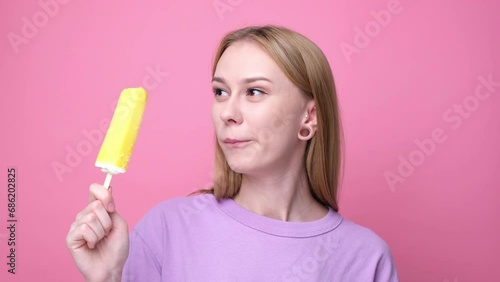 Happy young woman eating yellow ice cream on pink background. Concept of summer and sweet cold desserts. photo