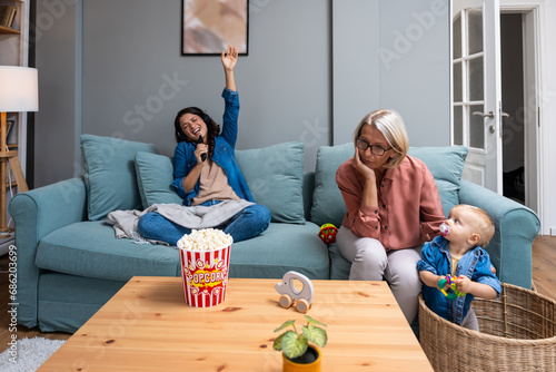 Young irresponsible mother watch TV and eat popcorn while her mother child grandmother looks after the baby. Carefree mom pays no attention to her child who is being looked after by older babysitter photo