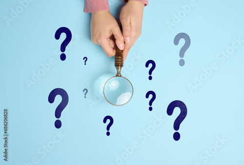 Female hand holds a wooden magnifying glass and question marks on a blue background. The concept of finding an answer to questions