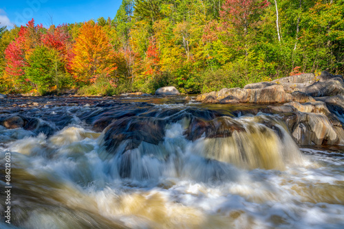 Vibrant autumn leaves on the riverbank with cascading water combine to create a symphony of nature's beauty.