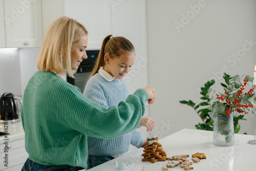 Happy mother and cute girl daughter decorating Christmas gingerbread cookies after baking while standing in cozy kitchen at home with Christmas decorations. photo
