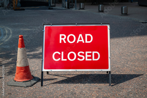 red road closed sign in the street with a traffic cone.