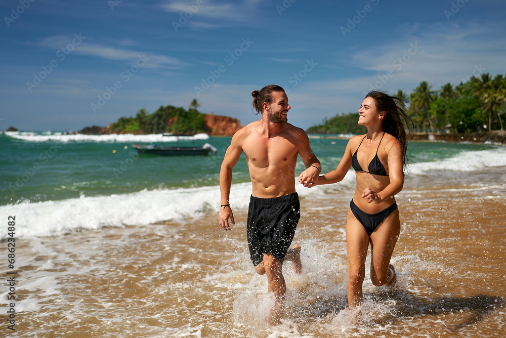 Joyful couple runs on tropical beach. Fit man and slender woman enjoy active vacation, sprinting by sea. Athletic duo practices beach running, promotes wellness. Excited pair in swimwear share fun.