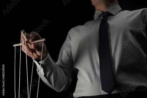 In the frame against a black background stands a man in a suit holding a device with ropes that hold a puppet. Depicts a businessman who manages someone, directs. Theatrical performance photo