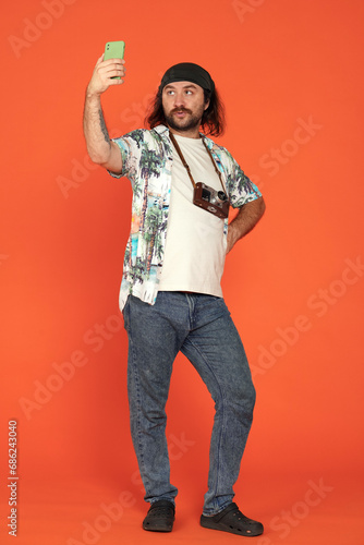 A male traveler with a retro camera uses a smartphone to take a selfie or speak via video call. Full length man in the studio on an orange background. Vertical shot.