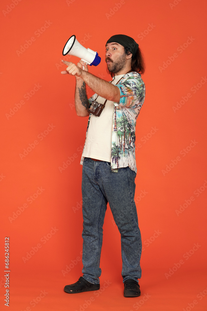 Surprised male traveler standing full length with megaphone, pointing at something with index finger. Man with camera around his neck in studio on orange background. Vertical shot. Concept of travel. Surprised male traveler standing full length with megaphone, pointing at something with index finger. Man with camera around his neck in studio on orange background. Vertical shot. Concept of travel.