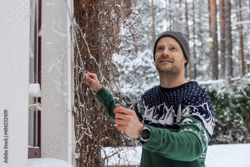 man decorating house exterior with christmas lights on snowy winter day