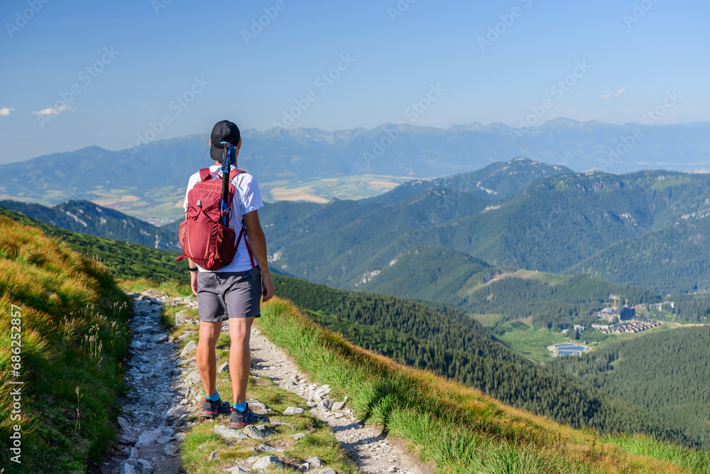 Polish Tatra Mountains, a young man in shorts and a cap with a mountain backpack stands on a mountain trail and looks at the valley with mountain peaks on a summer sunny day.