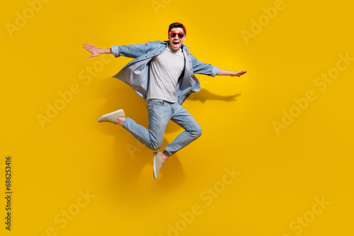 Full length photo of excited cheerful guy wear denim jacket dark glasses jumping high isolated yellow color background photo