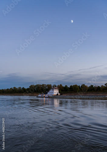 Freight barges passing over the calm waters of the Mississippi river at dusk with the moon high in the sky