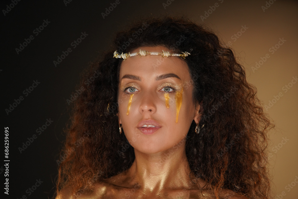 Portrait of female model in studio. Appealing girl wearing tiara and stylish makeup close up, body covered in golden paint with golden tears. Portrait of female model in studio. Appealing girl wearing tiara and stylish makeup close up, body covered in golden paint with golden tears.