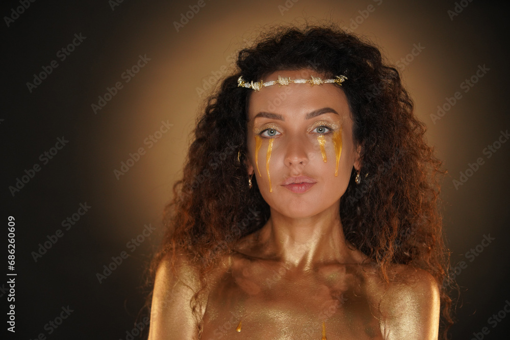 Portrait of female model in studio. Attractive girl wearing tiara and bright makeup shoulders area covered in golden paint, looking at the camera. Portrait of female model in studio. Attractive girl wearing tiara and bright makeup shoulders area covered in golden paint, looking at the camera.