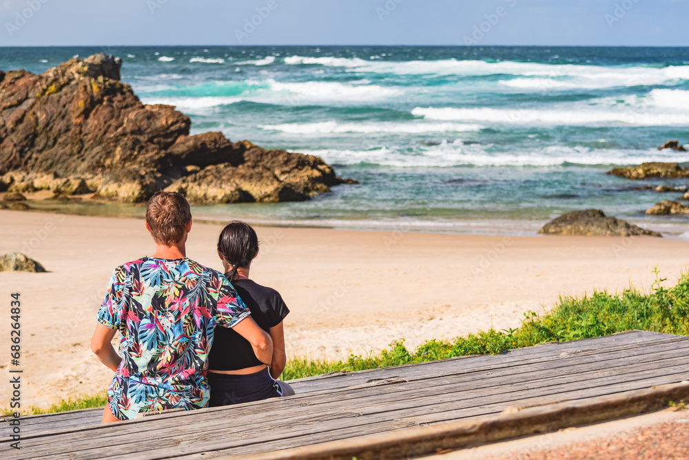 Young couple of embraced people sitting on a wooden bench at the shore with a wide beach, they look at the blue ocean and rocks at the shore, seaside landscape on a summer sunny day.