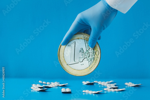 A hand in a blue medical glove holds a large 1 euro coin over scattered white tablets in blister packs on a blue background. Money for medicine, money for drugs, money for expensive treatment photo