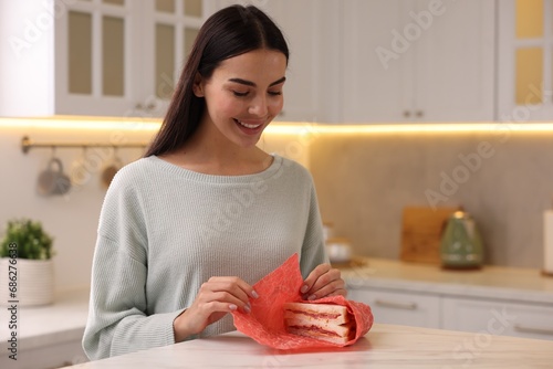 Happy woman packing sandwich into beeswax food wrap at table in kitchen photo
