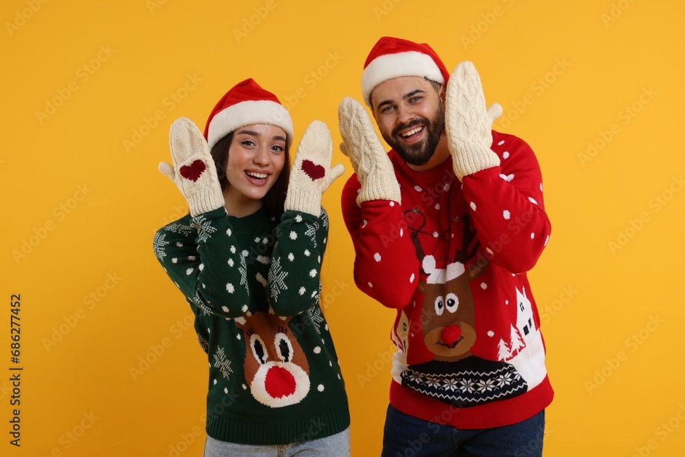 Young couple in Christmas sweaters, Santa hats and knitted mittens on orange background Young couple in Christmas sweaters, Santa hats and knitted mittens on orange background