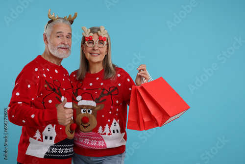 Senior couple in Christmas sweaters, reindeer headband and party glasses with shopping bags on light blue background. Space for text