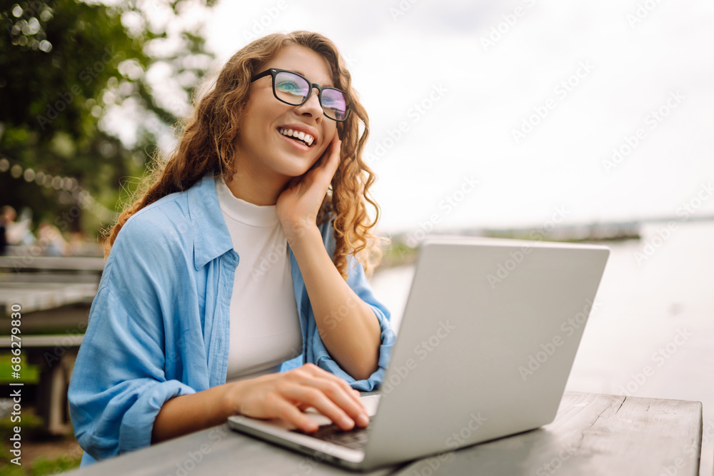 Portrait of a young woman in a blue shirt with a laptop at a table in an outdoor cafe near a lake. Curly woman freelancer works on a laptop outdoors. Freelance concept, nature. Portrait of a young woman in a blue shirt with a laptop at a table in an outdoor cafe near a lake. Curly woman freelancer works on a laptop outdoors. Freelance concept, nature.